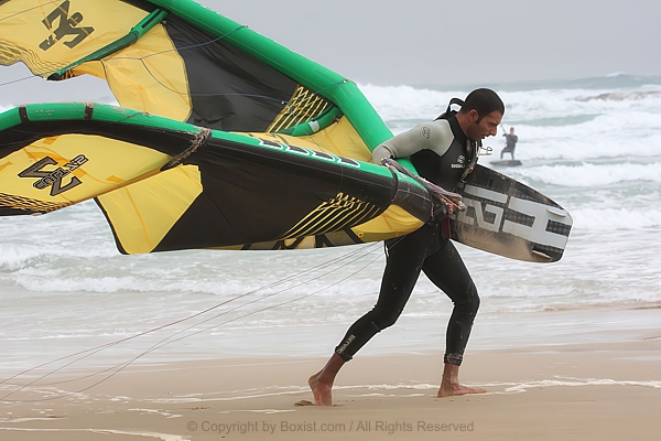 Man Carrying Kiteboarding Gear Along Shoreline Against Wind