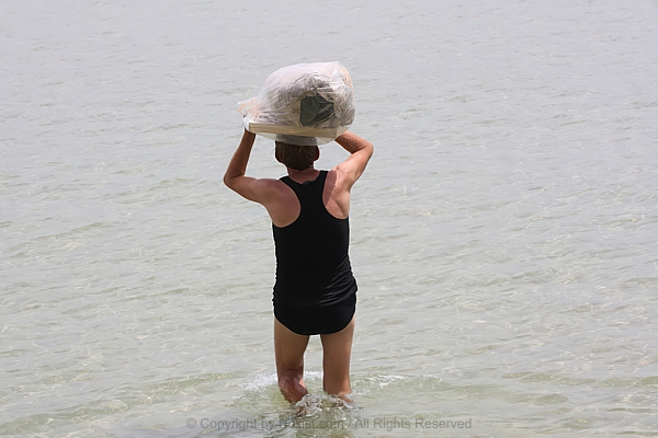 Person Wading Through Shallow Sea With Belongings In Bag On Head